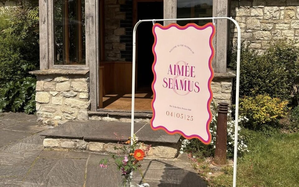 A wedding welcome sign stands outside a rustic stone building with a red-tiled roof and wooden-framed entrance. The pink sign with red wavy border reads, “Welcome to the wedding of Aimée & Seamus, The Tithe Barn, Petersham Hotel, 04/05/25.” A glass vase with colourful flowers sits at the base of the sign.