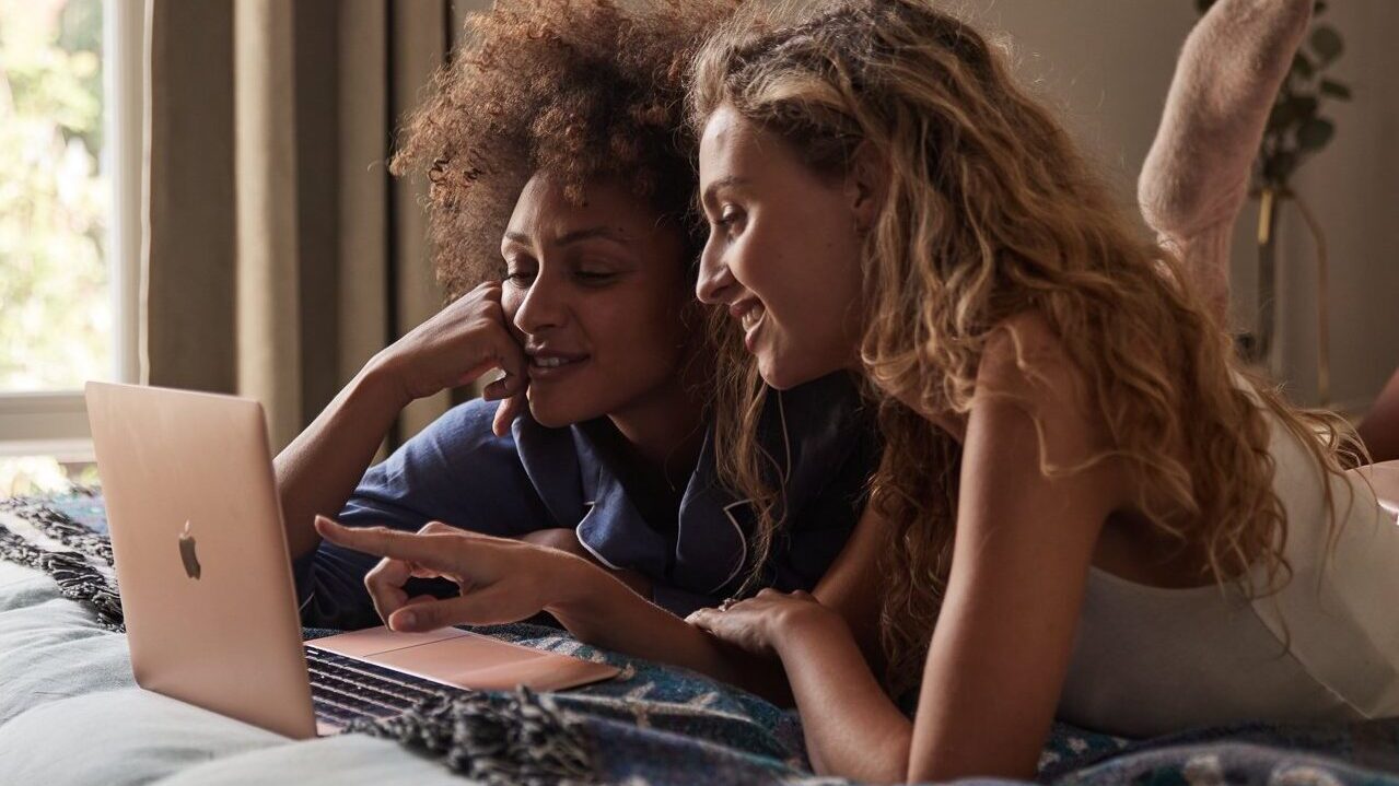 Two women sat on bed looking at apple macbook laptop 