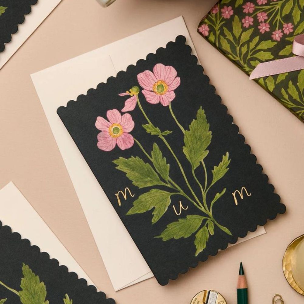 Dark green scalloped-edge card featuring pink illustrated flowers and gold lettering spelling “mum.”