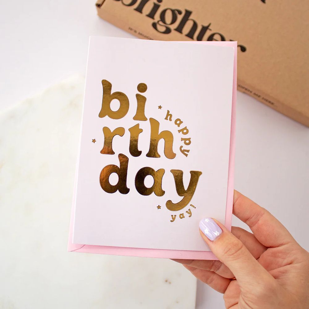 White card with shiny gold lettering arranged playfully to spell “happy birthday yay!”