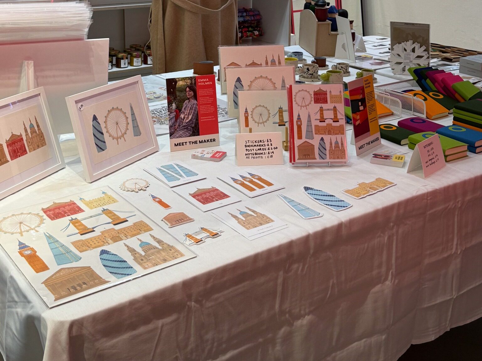 A neatly arranged craft market table displaying illustrated stationery and art prints featuring London landmarks such as Big Ben, the London Eye, Tower Bridge, St Paul’s Cathedral, and modern skyscrapers. Framed prints, sticker sheets, bookmarks, postcards, and small notebooks are laid out on a white tablecloth, with upright display stands and a “Meet the Maker” sign. Colorful notebooks are stacked to the right, and the overall scene looks like a designer’s stall at a craft or makers’ fair.