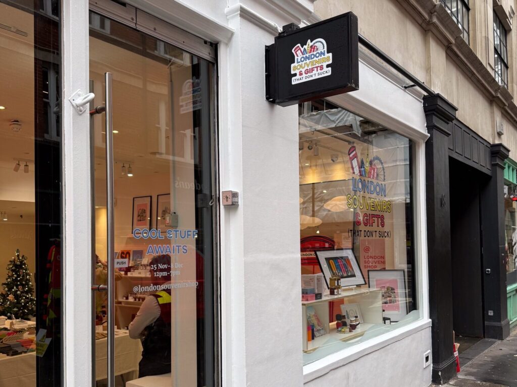 Exterior of a London souvenir shop with a white storefront and large glass windows. A black hanging sign reads ‘London Souvenirs & Gifts (That Don’t Suck).’ Through the windows, colorful displays of artwork and gifts are visible, along with a small decorated Christmas tree inside. Text on the door reads ‘Cool Stuff Awaits,’ with opening dates and the shop’s social handle.