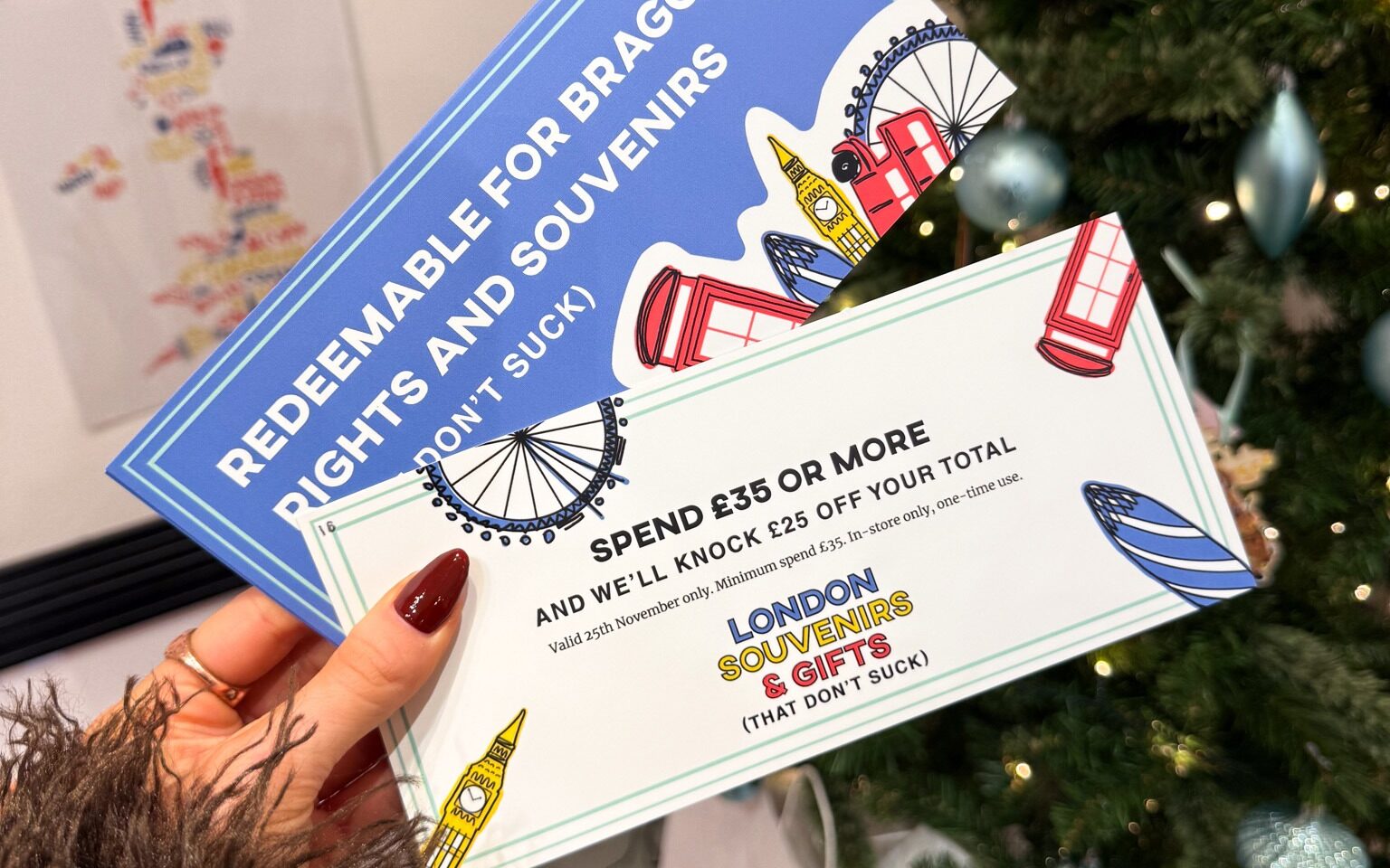 Close-up of a hand holding two illustrated vouchers for a London souvenir shop. The top blue voucher reads ‘Redeemable for Brag Rights and Souvenirs (That Don’t Suck).’ The front voucher says ‘Spend £35 or more and we’ll knock £25 off your total,’ with colorful illustrations of London icons like a red telephone box, Big Ben, and the London Eye. A decorated Christmas tree with lights and ornaments is blurred in the background.