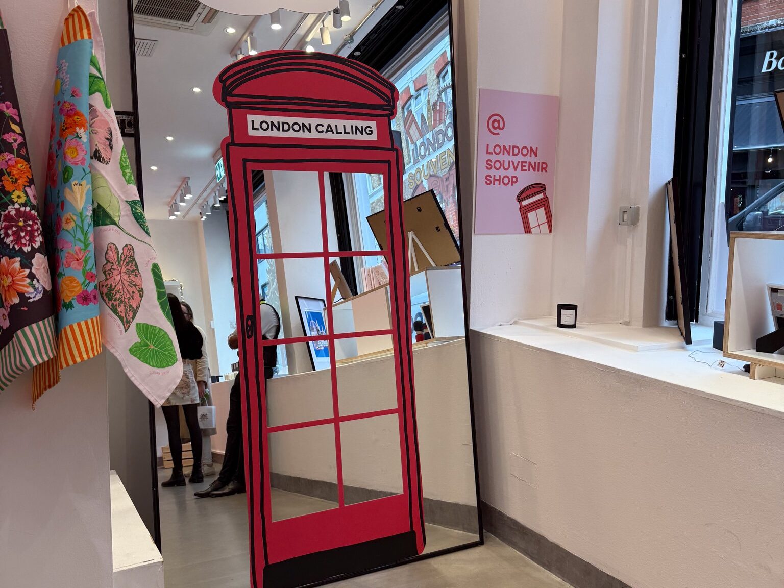 Interior of a London souvenir shop featuring a tall mirror shaped like a red British telephone box with the words ‘London Calling’ at the top. Scarves hang on the left, framed artwork and small displays sit on white shelves, and a pastel sign on the wall reads ‘London Souvenir Shop.’ Reflections of shoppers and the street outside are visible in the mirror.