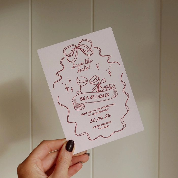 A hand holding a Save the Date with an illustrated design featuring bows, wine glasses and stars. 