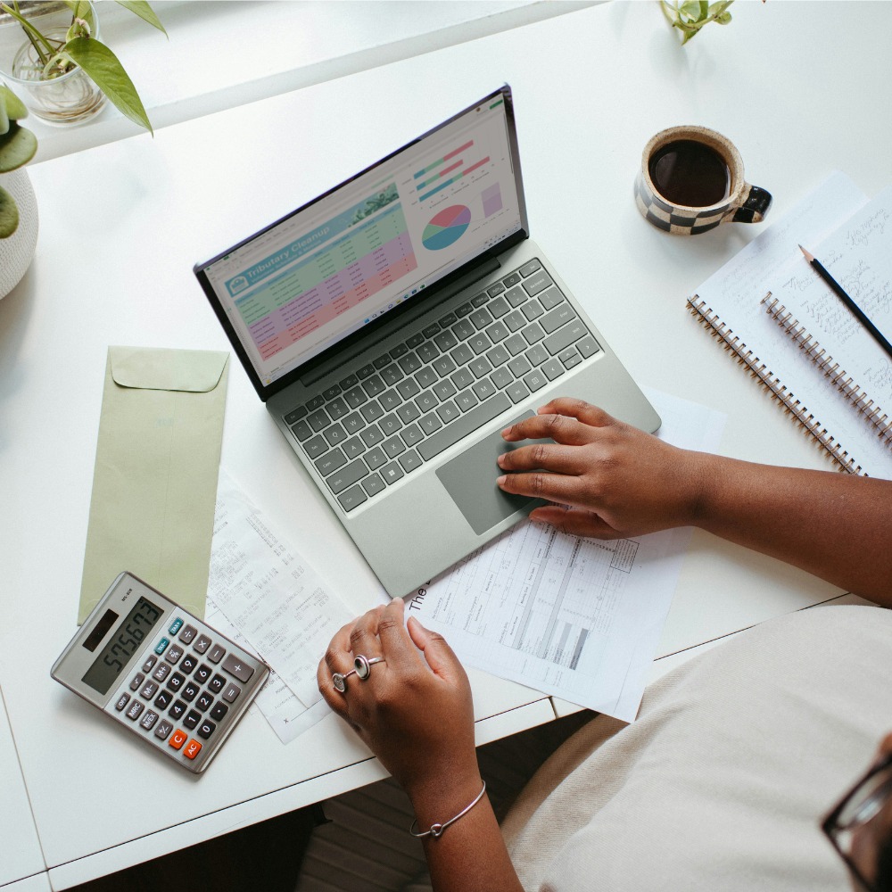 A photograph taken from above. It shows a woman's hands working on a laptop, she's looking at a budget and is surrounded by a calculator, paperwork, a notebook and a coffee.