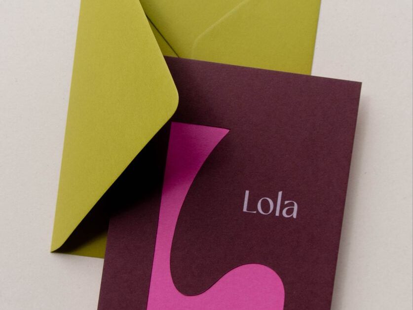 A minimalist stationery set featuring a deep plum-colored card with a large, abstract pink "L" and the name "Lola" in small white serif text. The card rests against an open, chartreuse green envelope on a light cream background.