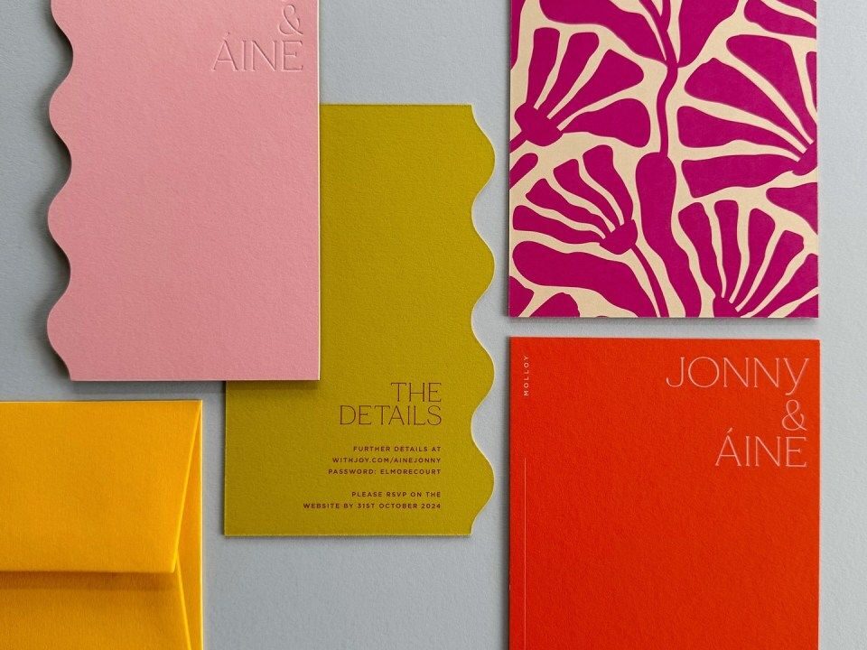 A flat-lay photograph of a modern, colorful wedding invitation suite arranged on a neutral grey background. The suite features a vibrant color palette of pink, mustard yellow, olive green, and bright orange.