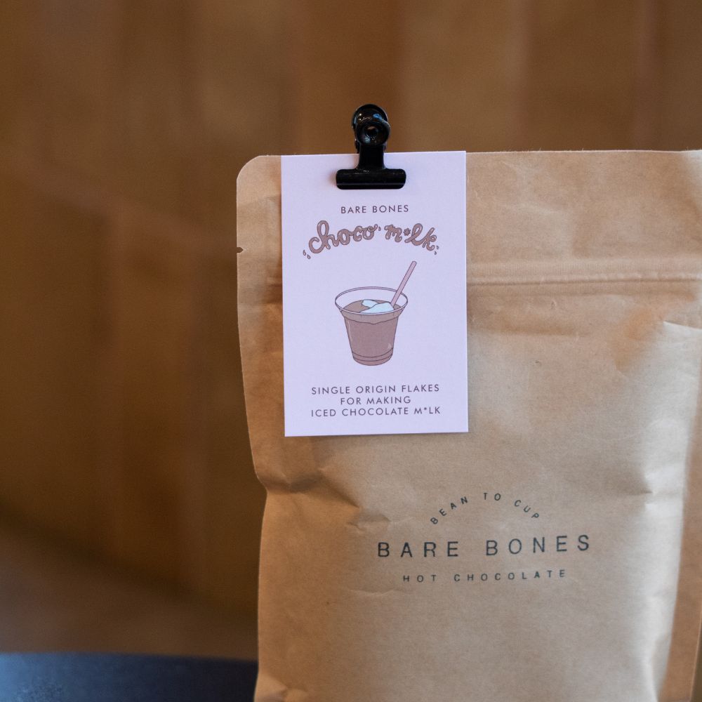 A Business Card for Bare Bones Chocolate Milk, which features an illustration of a cup of chocolate milk. It is attached to a bag of Bare Bones Hot Chocolate.