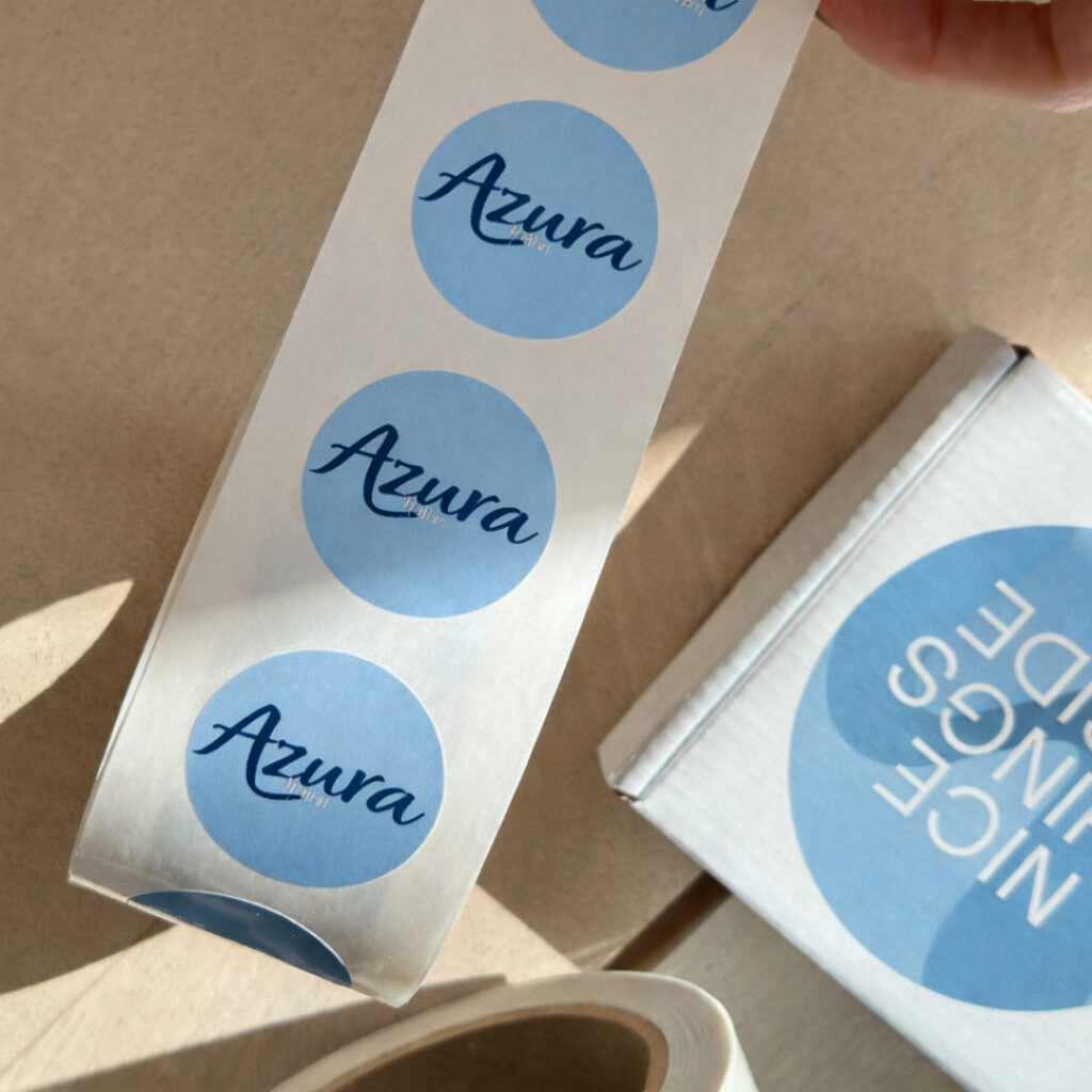 A roll of light blue circular stickers featuring the word "Azura" in a navy blue script font. The roll is held above a white cardboard box that has a matching blue sticker reading "NICE THINGS INSIDE" in white uppercase letters.