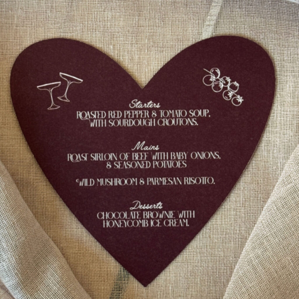 A heart-shaped wedding menu card in a deep plum or burgundy color, resting on a textured beige fabric. The menu features white text with decorative line drawings of cocktail glasses and a sprig of berries. The courses listed are Starters (Roasted Red Pepper & Tomato Soup), Mains (Roast Sirloin of Beef or Wild Mushroom Risotto), and Desserts (Chocolate Brownie).