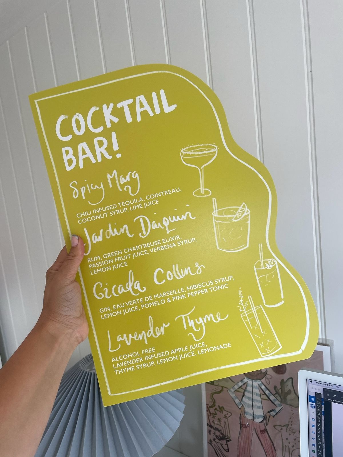 A hand holds a large, uniquely shaped cocktail menu card in a bright chartreuse or lime green color. The menu is titled "COCKTAIL BAR!" in bold white letters and features a wavy, die-cut right edge. It lists four drinks—Spicy Marg, Jardin Daiquiri, Cicada Collins, and an alcohol-free Lavender Thyme—accompanied by simple white line illustrations of different cocktail glasses.