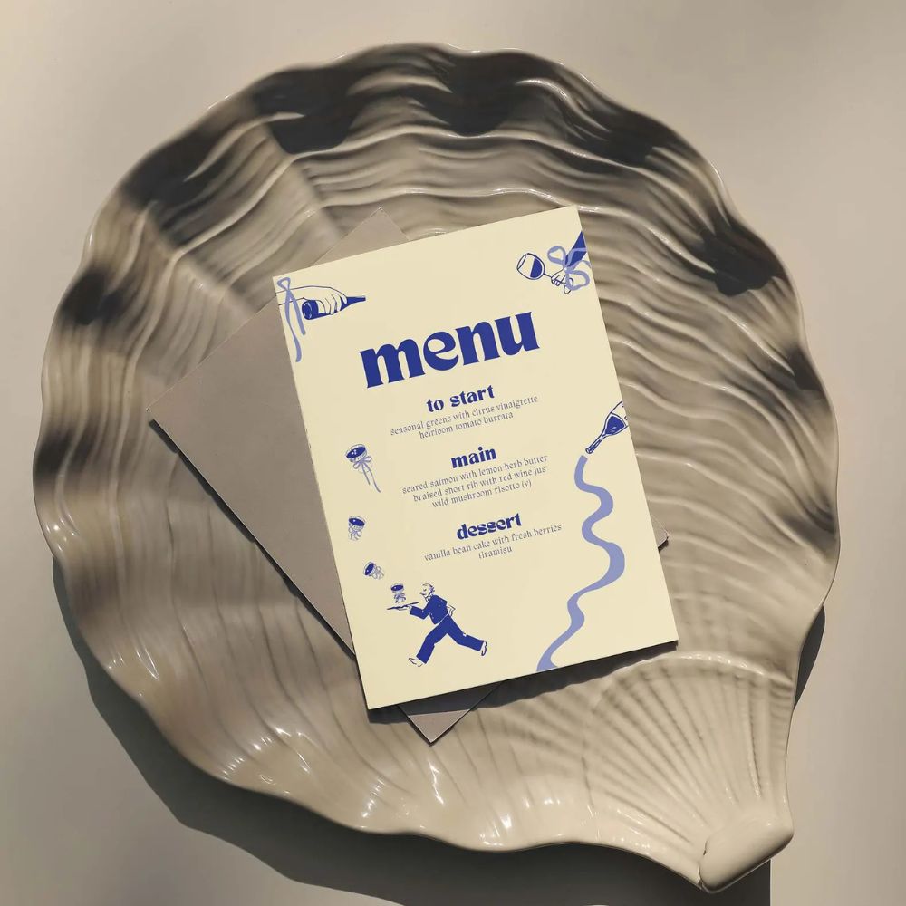 A cream Wedding Menu with blue text and images, photographed on a shell shaped plate. 