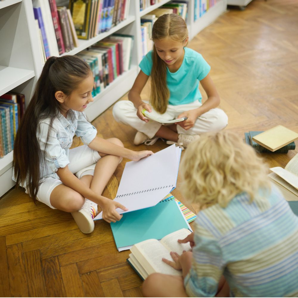 Three young children sat on the floor by some bookshelves, all reading books. 