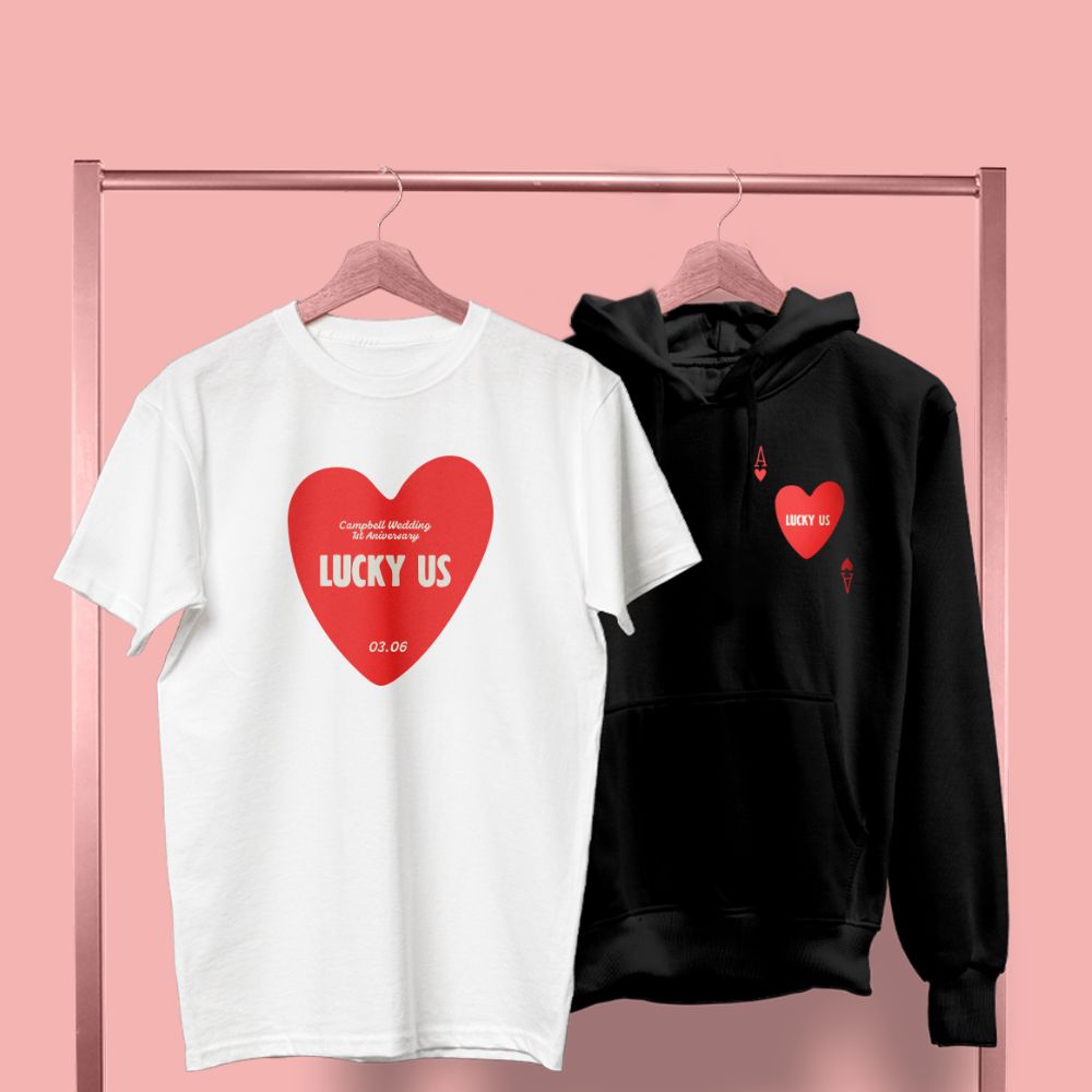 A white T-Shirt and a black hoodie hanging side by side, both featuring a red heart design that reads "lucky us" in white text. 