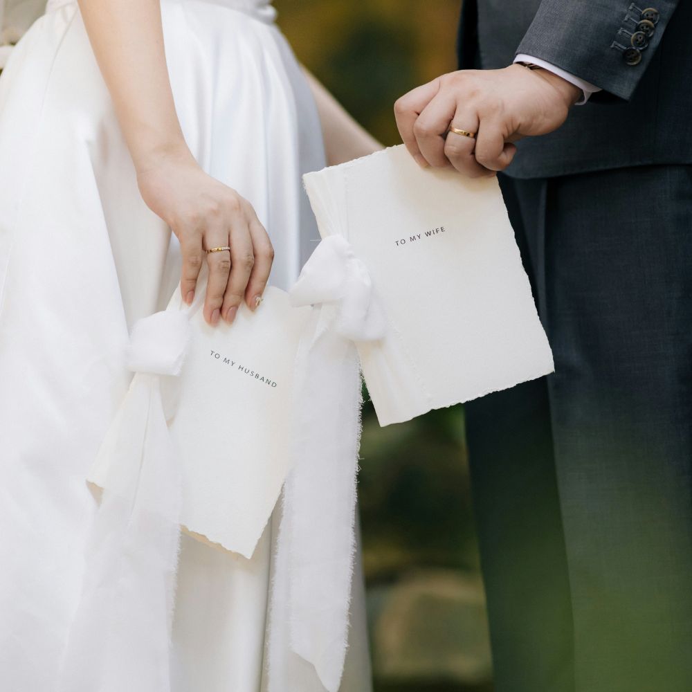 The hands of a couple getting married, each holding vows. They say "to my husband" and "to my wife". 