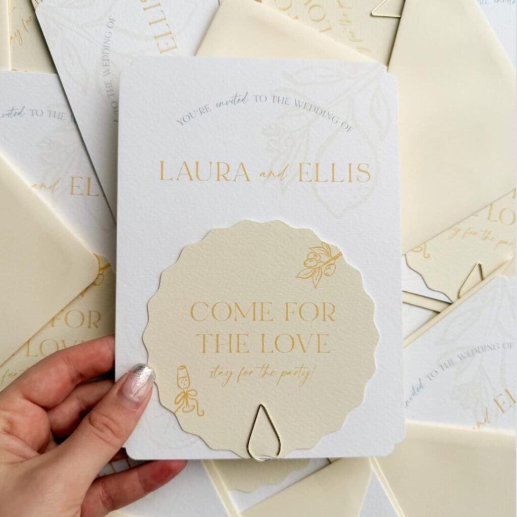 A person’s hand holds a wedding invitation featuring a soft butter yellow and white colour scheme, set against a background of scattered matching cards and envelopes. The main invitation is a white textured rectangle with rounded corners, featuring the names Laura and Ellis in yellow capital letters. Attached to the front with a gold teardrop-shaped paperclip is a smaller, scalloped-edge card in pale yellow. This card displays the phrase "COME FOR THE LOVE" in bold yellow text, followed by "stay for the party!" in a gold script. Subtle line illustrations of a lemon branch and a champagne glass decorate the stationery, creating a fresh, spring-inspired aesthetic.