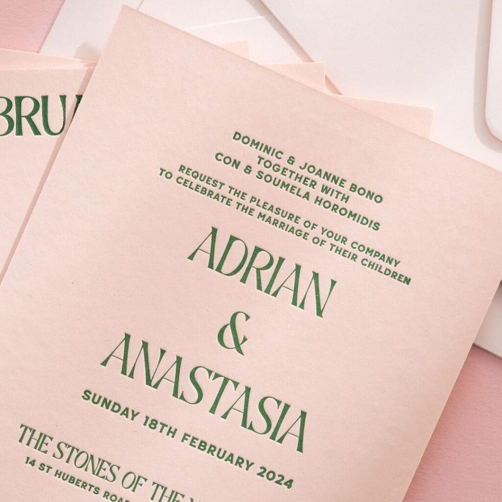 A close-up shot shows a stack of peach-coloured wedding invitations with elegant dark green lettering. The top card features the names Adrian & Anastasia in a large, classic serif font. Above their names, the text invites guests to celebrate their marriage on behalf of their parents. The date Sunday 18th February 2024 is printed below the names, followed by the venue address, The Stones of the Yarra Valley. The invitations are set against a soft pink background, creating a warm and sophisticated colour palette.