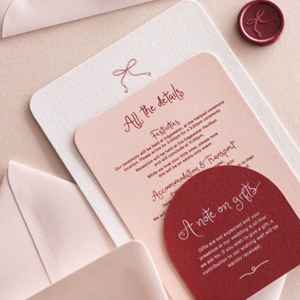 A tiered wedding invitation suite in rose pink and rouge red tones is arranged on a neutral background with two pale pink envelopes and a wax seal. The main cards have rounded corners and are layered on top of each other.
The largest card is off-white with a small red line drawing of a bow at the top. Layered over it is a dusty pink card titled "All the details" in red script, featuring information about the wedding festivities and transport. At the bottom right, a small rouge red card with an arched top is titled "A note on gifts" in white script, explaining the couple's wishing well. A matching rouge wax seal with an embossed bow sits in the top right corner, completing the romantic and sophisticated stationery set.