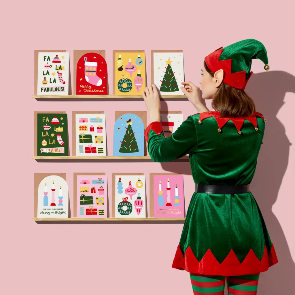A woman dressed as a Christmas elf displaying Christmas cards on wall shelves. 