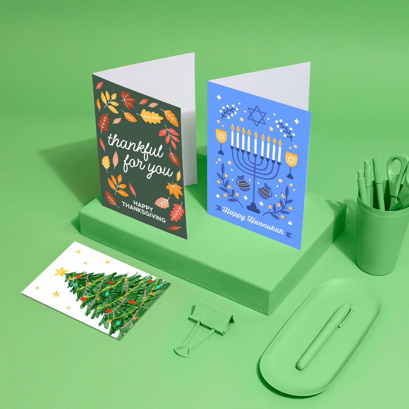 Three holiday cards, one for Thanksgiving with autumn leaves, one for Hanukkah with a menorah and dreidels, and one for Christmas featuring a decorated tree are displayed on a green background alongside green office supplies.