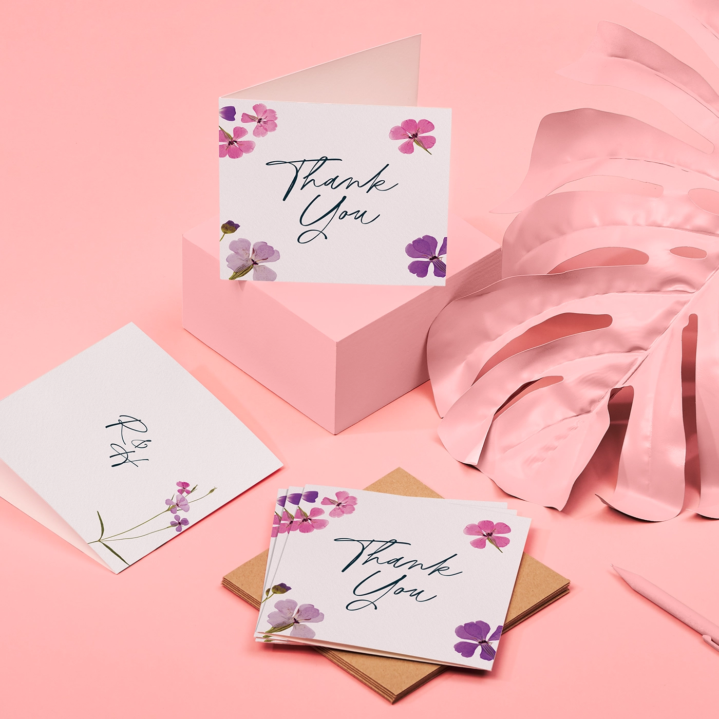 Several folded wedding thank you cards with a simple white background and a pink and purple floral design, on a soft pink background.