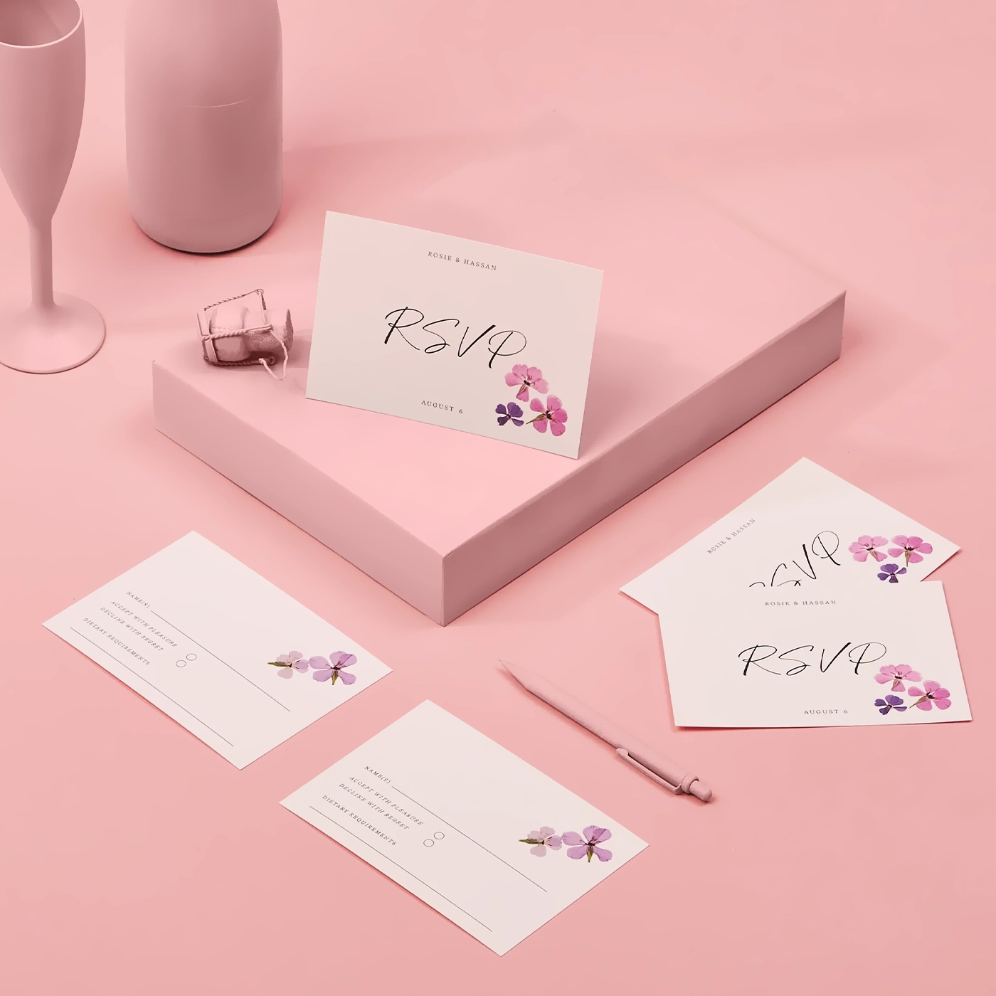 Several floral RSVP cards for Rosie and Hassan, decorated with purple and pink flowers, are arranged on a soft pink surface alongside a matching pink pen, box, and matte champagne flute and bottle.