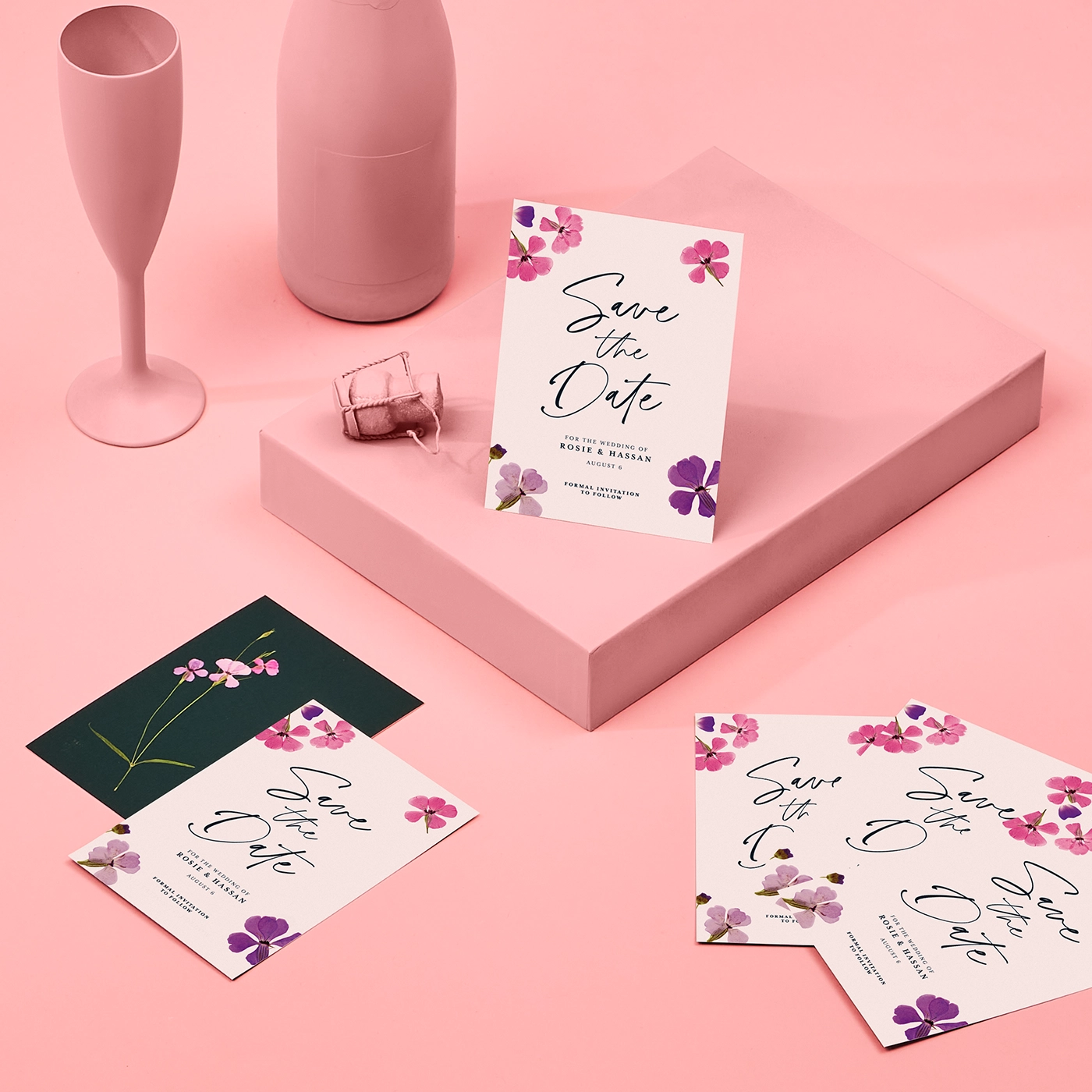 Several floral save the date cards decorated with purple and pink flowers, are arranged on a soft pink surface alongside a matching pink box, and matte champagne flute and bottle.