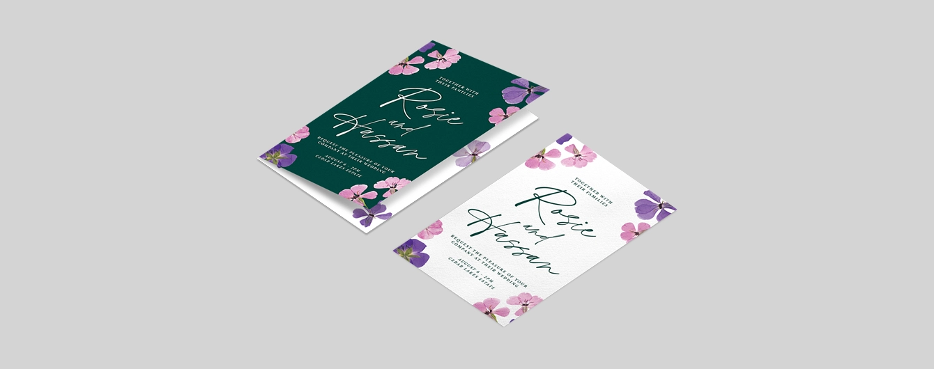 One flat and one folded wedding invitation with a green, white, pink and purple floral design, laid on a plain background.