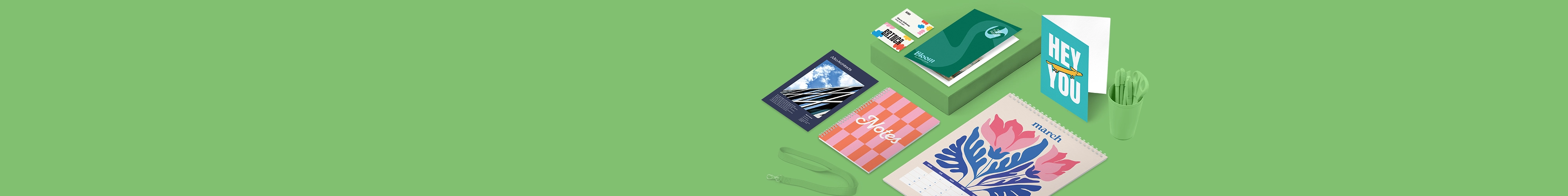 A selection of print products including greeting cards, a calendar, bookmarks and a notebook with pink, organge and blue colorful designs, laid on a green background featuring a pencil pot and lanyard.