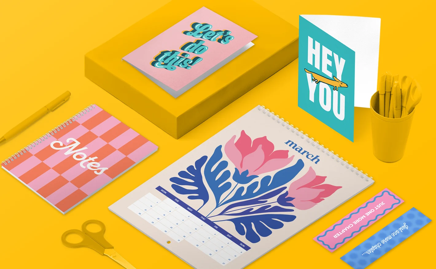 A selection of print products including greeting cards, a calendar, bookmarks and a notebook with pink, organge and blue colorful designs, laid on a yellow background featuring scissors and pencils.