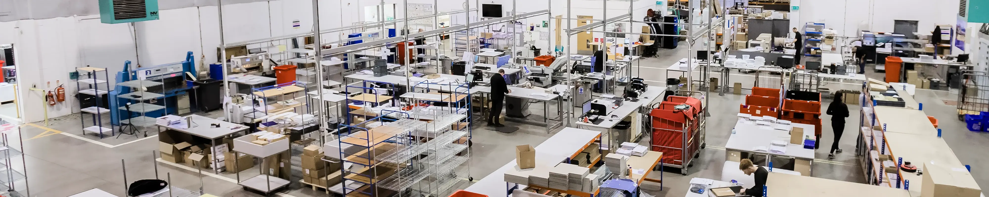 A large, brightly lit warehouse or production facility with multiple workstations, tables, and shelving units; several people are working on various tasks, surrounded by boxes, carts, and office equipment.