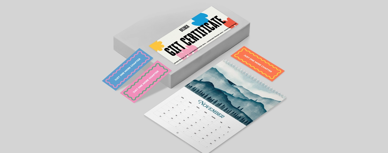 Flat lay of a white Gift Certificate featuring bold black text and colorful abstract shapes. Next to the certificate are three colorful bookmarks, each reading 'Just One More Chapter' in white text—one blue, one pink, and one orange. Below is a spiral-bound calendar showing the month of November with an illustrated blue watercolor mountain landscape.