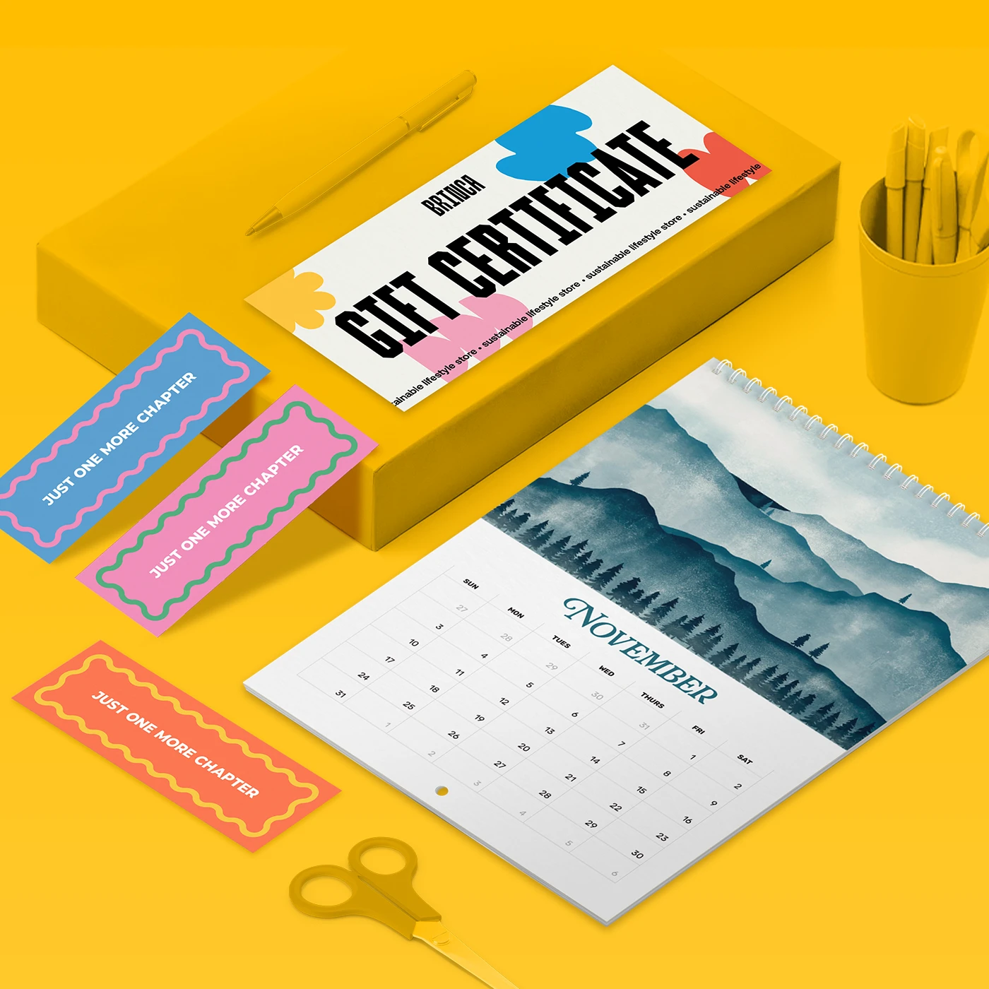 Flat lay of a white Gift Certificate featuring bold black text and colorful abstract shapes. Next to the certificate are three colorful bookmarks, each reading 'Just One More Chapter' in white text—one blue, one pink, and one orange. Below is a spiral-bound calendar showing the month of November with an illustrated blue watercolor mountain landscape.