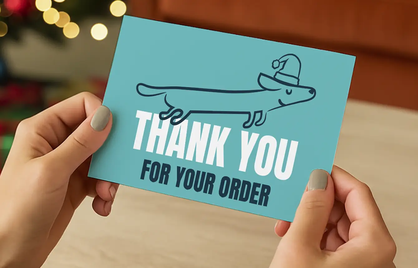 Someone holding a blue printed Postcard with text reading 'Thank You For Your Order'