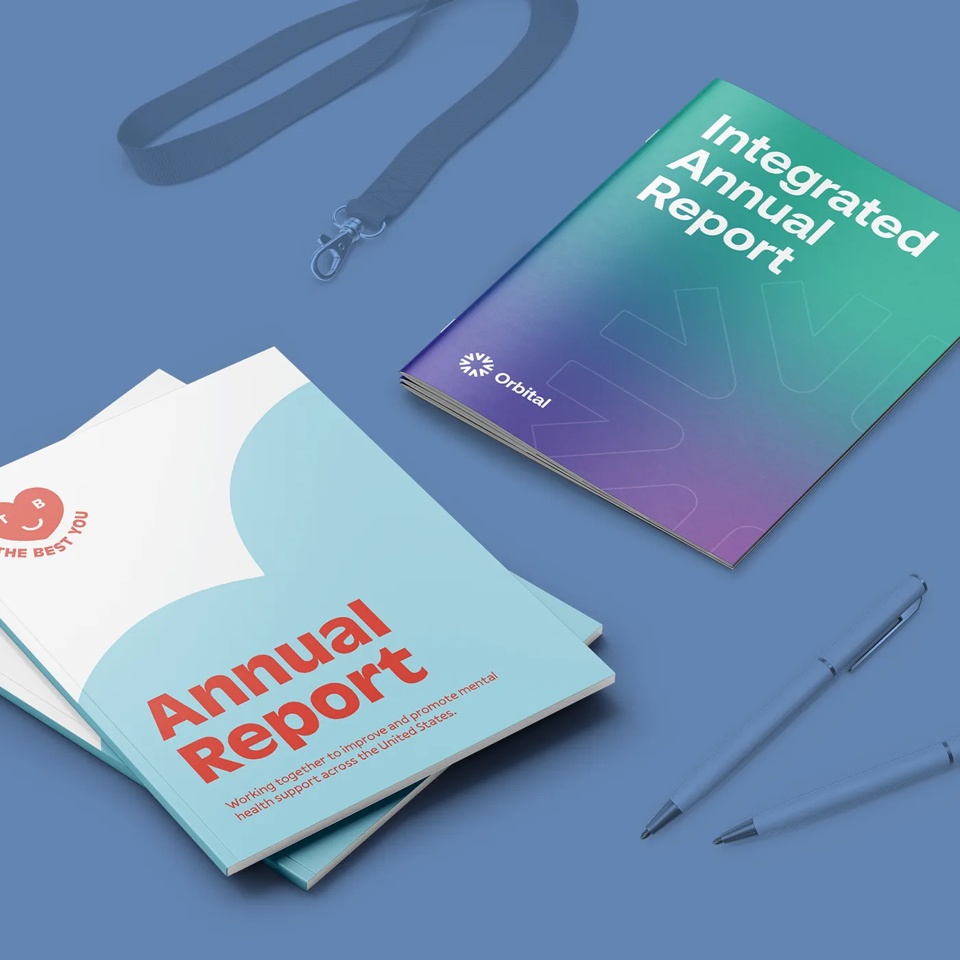 A blue surface displays an "Integrated Annual Report" with a teal-to-purple gradient cover alongside a stack of "Annual Reports" for a mental health organization, two blue pens, and a blue lanyard.