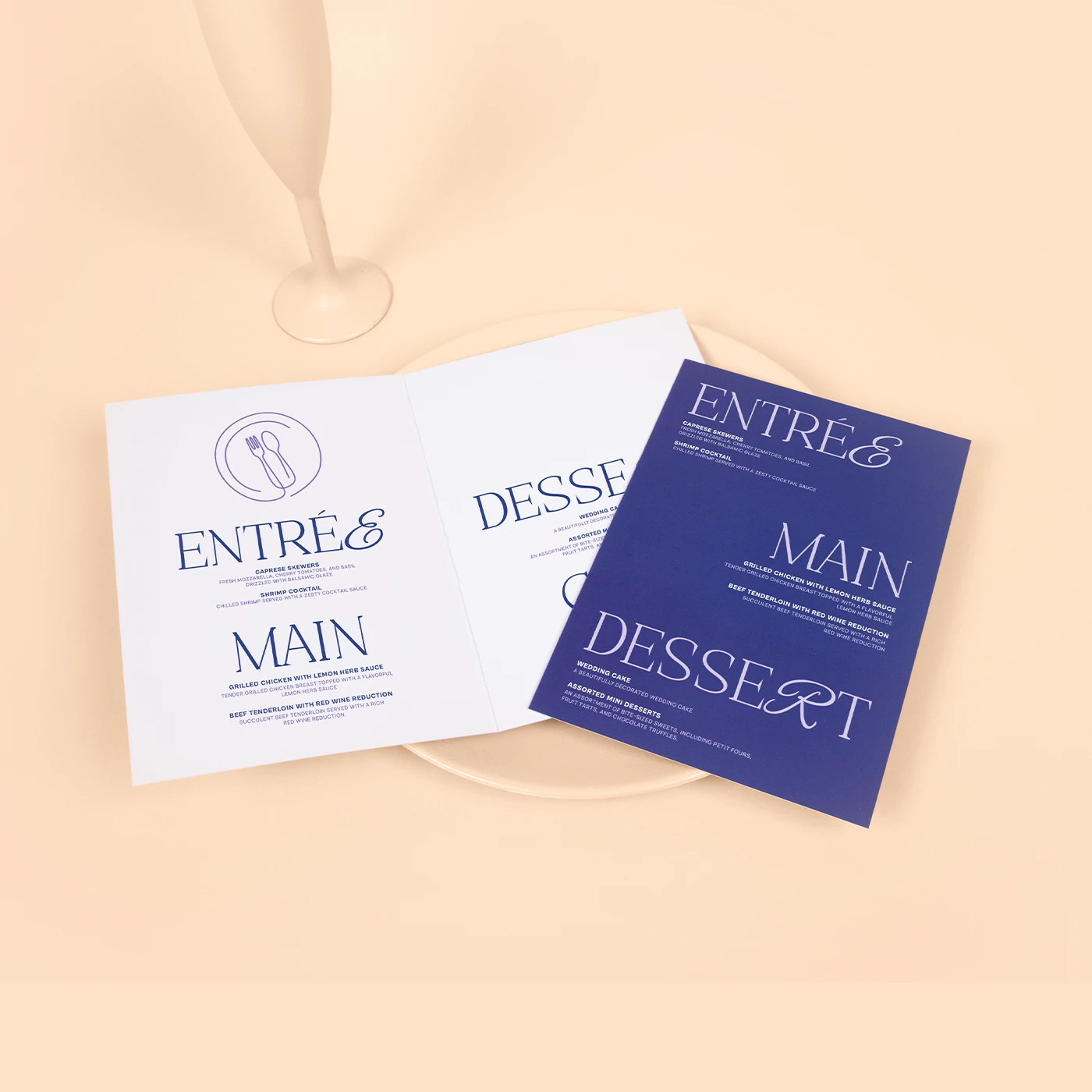 A bifold wedding menu is displayed against a pale peach background, featuring two versions: one with dark blue text on white cardstock and another with white text on dark blue cardstock. The menu sections—"ENTRÉE," "MAIN," and "DESSERT"—are printed in a large, elegant serif font, with detailed dish descriptions listed underneath each heading.