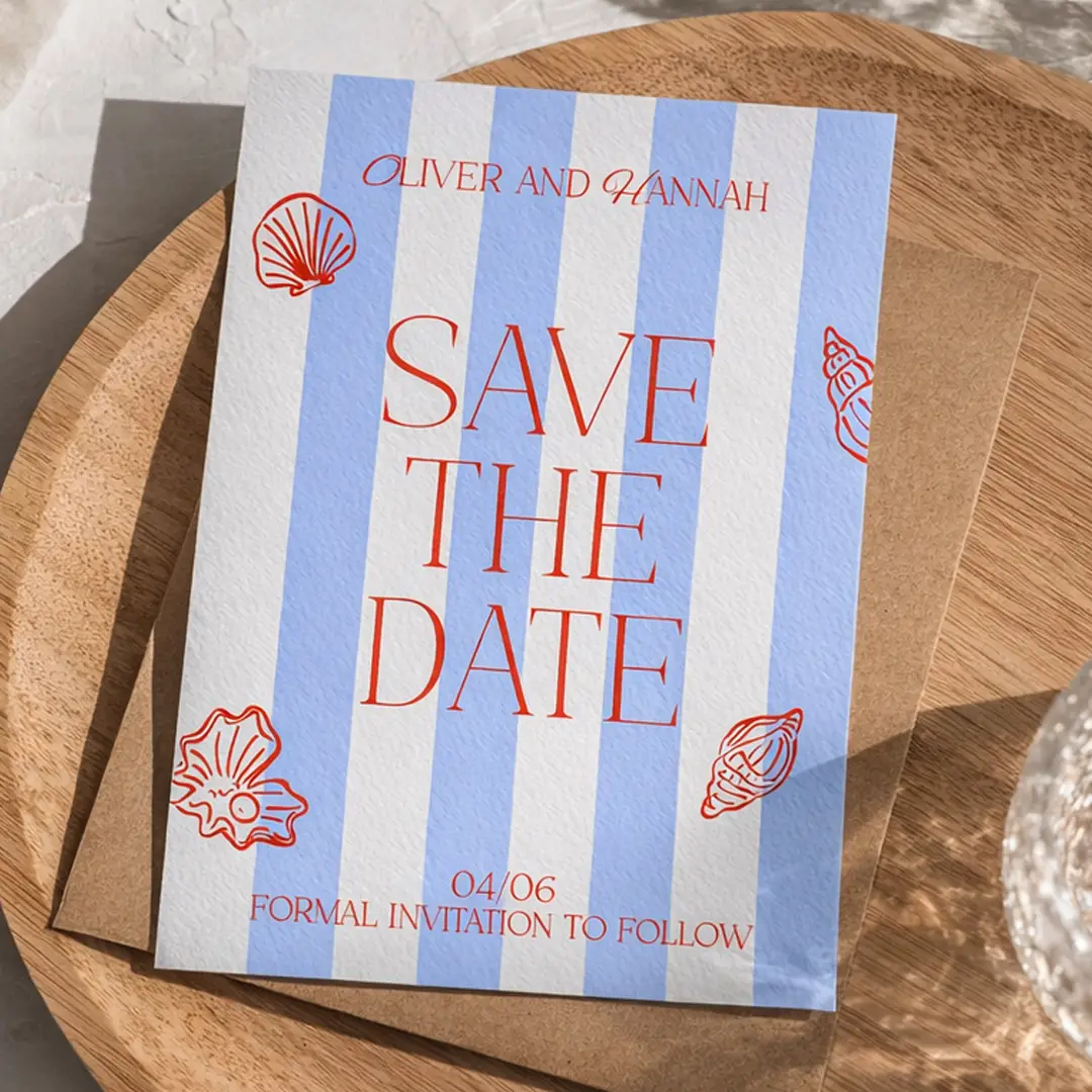 A nautical-themed "Save the Date" card for Oliver and Hannah rests on a brown envelope atop a wooden surface. The card features light blue and white vertical stripes, bold red serif text, and delicate red seashell illustrations in the corners, with the date "04/06" and the note "Formal invitation to follow" printed at the bottom.