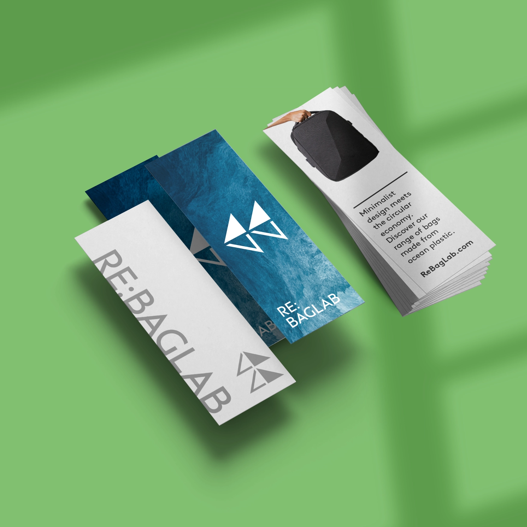 A stack of narrow, vertical mini business cards for "RE:BAGLAB" is displayed on a green surface with soft shadows. The cards feature three distinct designs: one with a minimalist white background and grey text, one with a deep blue textured ocean pattern and a white logo, and one showing a black backpack with descriptive text about circular economy products.