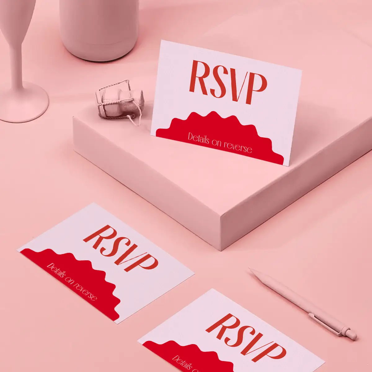 A white RSVP card with bold red text and a wavy red border at the bottom sits propped on a pink pedestal alongside two matching cards. The scene is styled on a monochromatic pink background with a pink pen and a champagne cork.