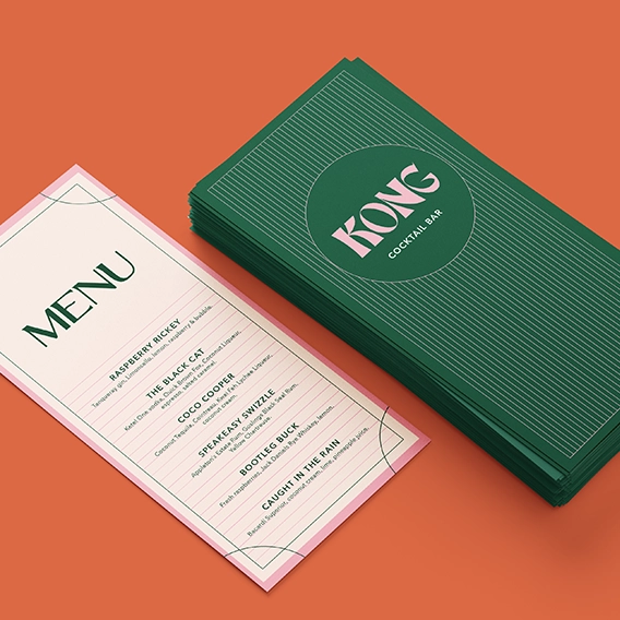 A narrow, vertical menu for "KONG Cocktail Bar" featuring a cream background and green text lies next to a stack of dark green flyers with the same brand name in bold pink and white. The menu lists several specialty drinks, while the green flyers feature a pattern of thin, horizontal white lines across the front.