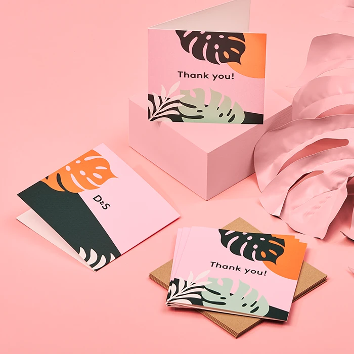 Gemini said A collection of square "Thank You" cards for "D&S" featuring a tropical monstera leaf design in pink, green, orange, and black. One card is propped open on a pink pedestal, while others are scattered on a pink surface alongside brown envelopes and a large decorative pink leaf.