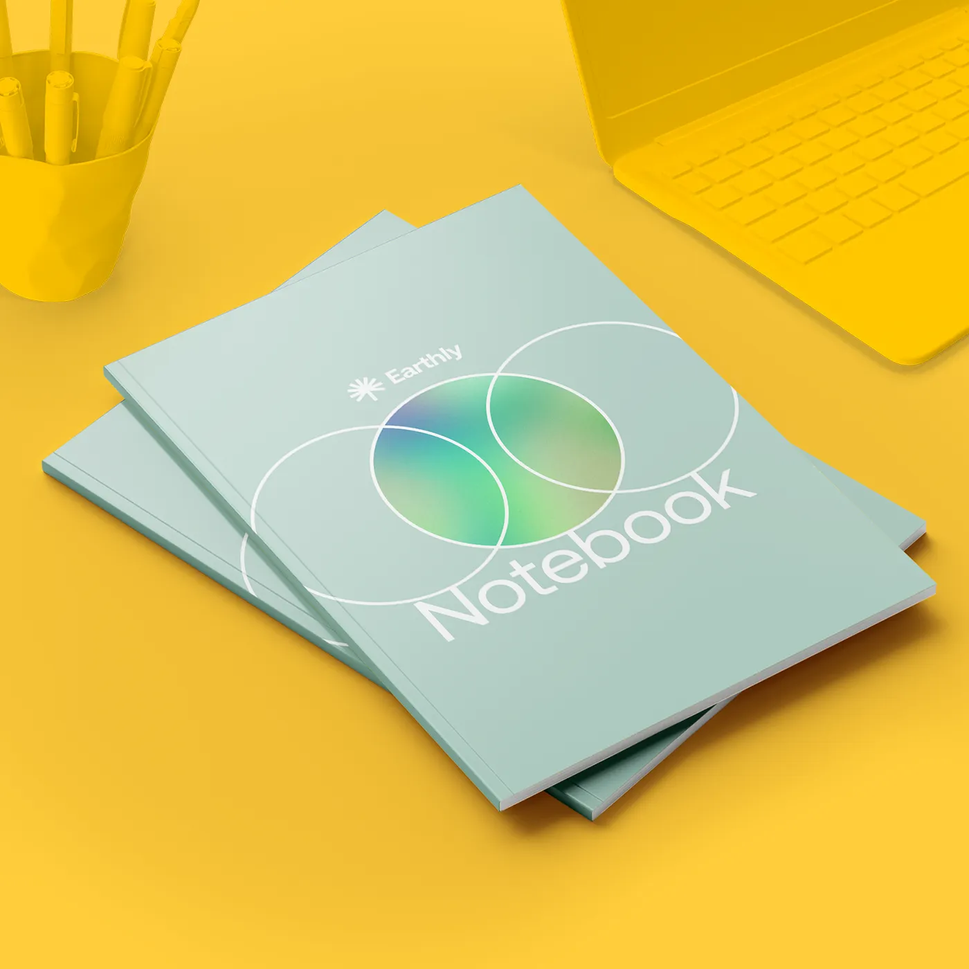 Two stacked blue Perfect Bound Notebooks with a circular gradient design are displayed on a vibrant yellow desk next to a laptop and pen holder.