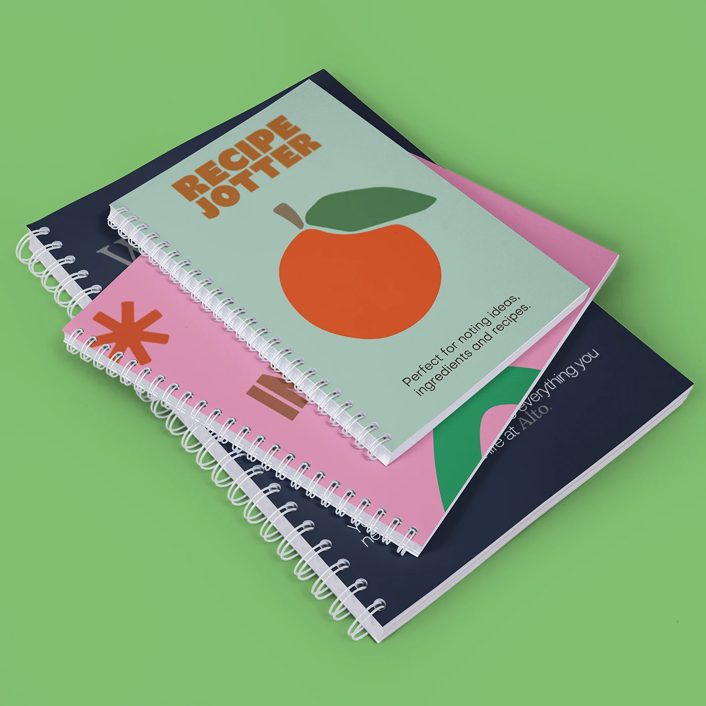 Three stacked Spiral Bound Notebooks: the top one is light green with an orange illustration and the title ‘Recipe Jotter,’ the second has a pink cover, and the third has a dark blue cover.