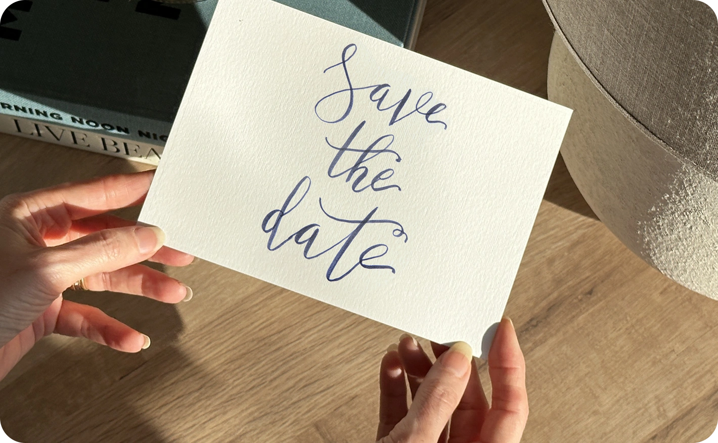 A person holding a white Save the Date card with cursive blue lettering. The background shows a modern interior with a wooden floor and soft shadows.