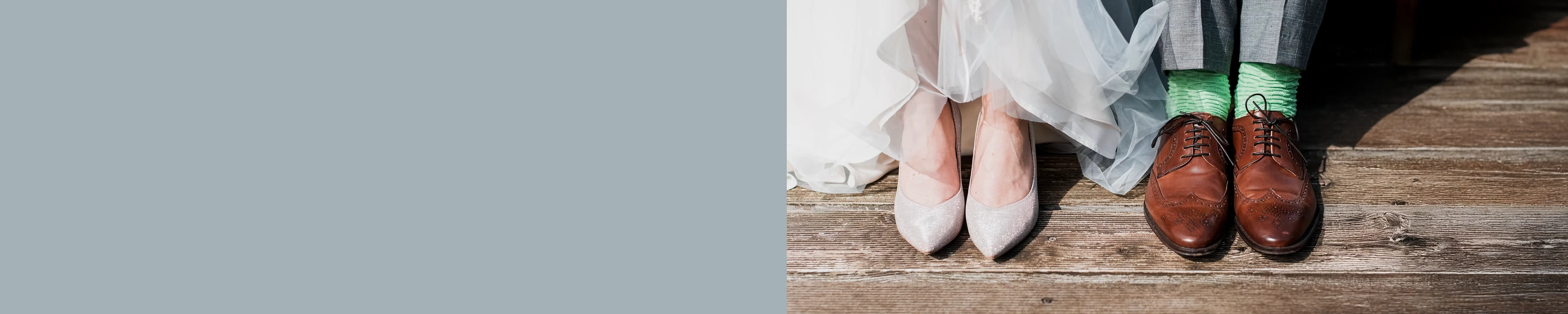 A close-up, low-angle shot of a bride and groom's feet standing on a rustic wooden deck. The bride wears shimmering silver pointed-toe heels beneath the hem of her white tulle gown. Beside her, the groom wears classic brown leather wingtip dress shoes with vibrant lime green textured socks and grey trousers. The left side of the banner features a solid slate blue color block for text overlay.
