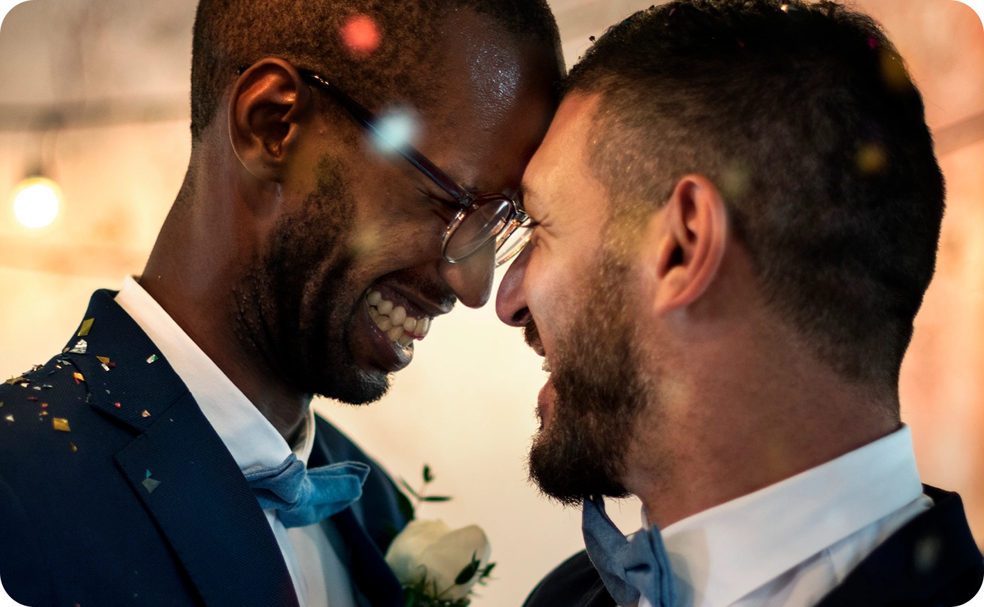 A candid, close-up shot of two grooms laughing together as confetti falls at their wedding reception. The scene is lit with warm ambient lighting, capturing a celebratory and romantic atmosphere.