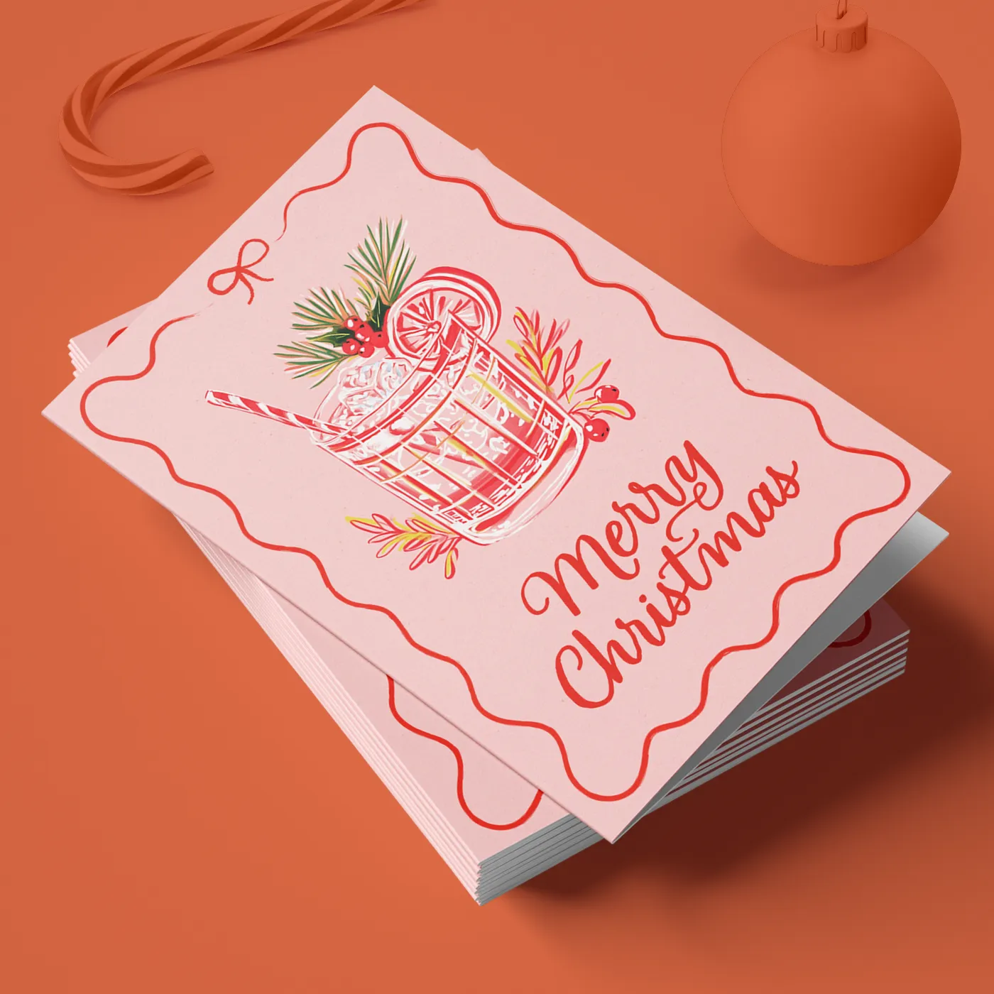 A stack of pink holiday cards featuring a whimsical illustration of a festive cocktail garnished with evergreen sprigs and berries. The words "Merry Christmas" are written in red script, and the cards are set against a monochromatic orange background with a matching candy cane and ornament.