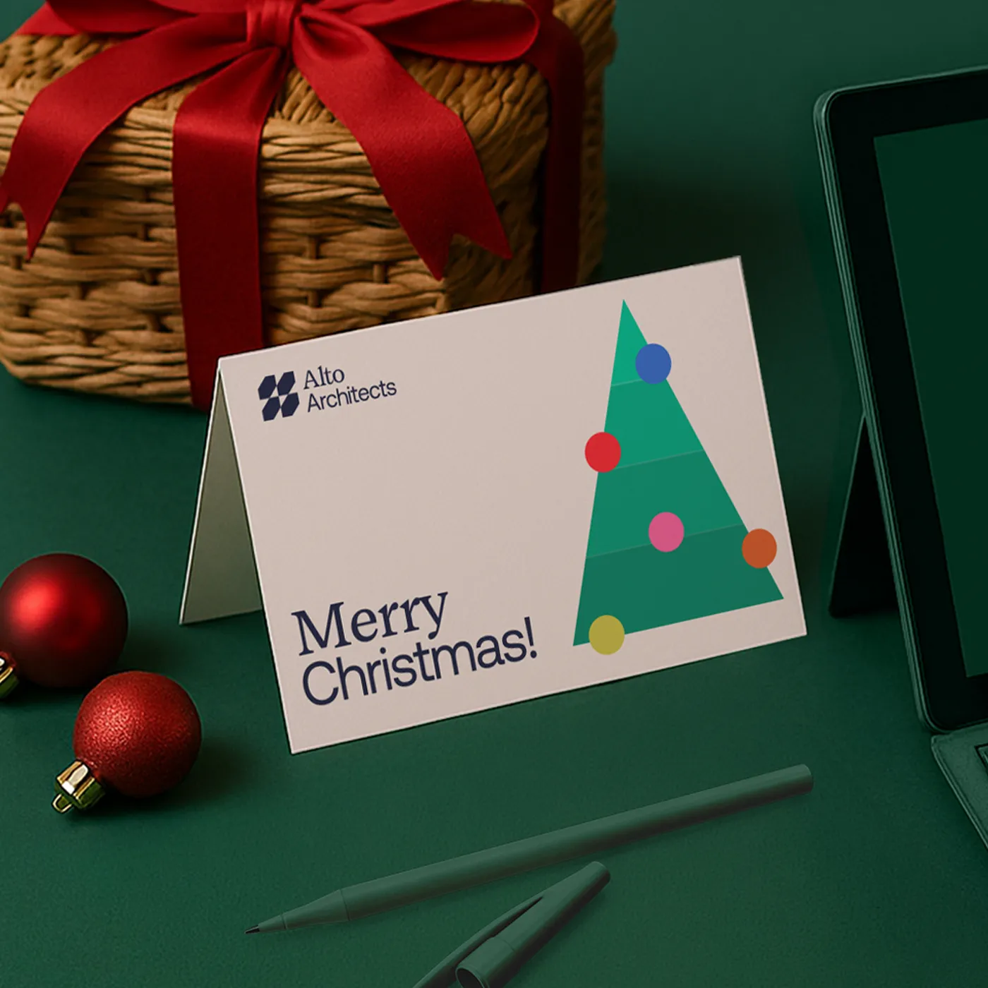 A professional "Merry Christmas" card from Alto Architects featuring a minimalist, geometric Christmas tree with colorful ornaments. The card is displayed on a dark green surface alongside a wicker gift basket with a red bow, red ornaments, and green office supplies.