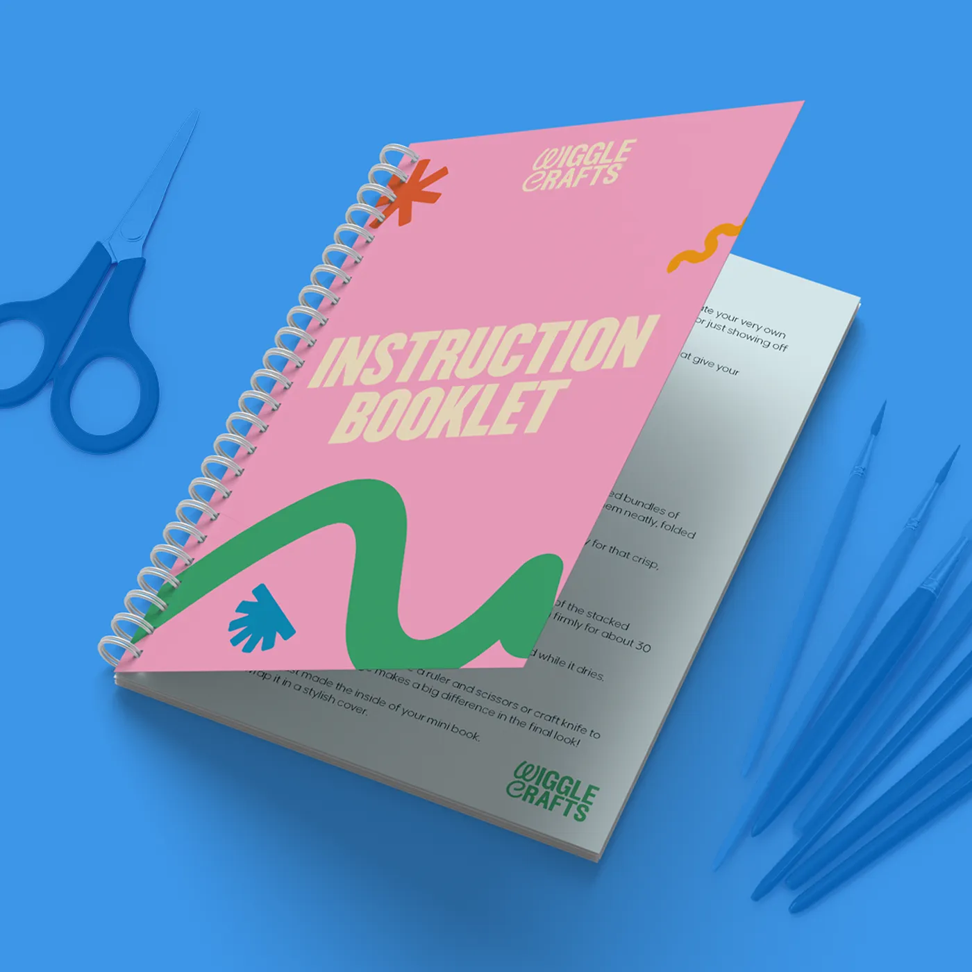 Gemini said A pink, spiral-bound "INSTRUCTION BOOKLET" for Wiggle Crafts is shown slightly open on a bright blue background. It is accompanied by matching blue craft supplies, including a pair of scissors and several paintbrushes.